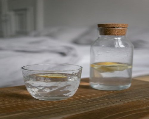 A transparent glass bottle filled with clean drinking water and a fresh slice of lemon inside on a wooden desk
